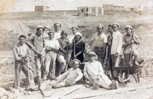 1927, קבוצת פועלים יוצאי "גדוד העבודה" בעבודות ציבוריות יזומות (הכשרת קרקע) בשכונת בורוכוב להתמודדות עם האבטלה הקשה, תוצר המשבר הכלכלי החמור שפקד את הארץ בשלהי תקופת העליה הרביעית. עומד משמאל, מקוש בידיו, ממנהיגי קבוצות העבודה בגדוד, יצחק לוי. שתי הבנות (אחיות ?) במרכז, אף שלצערנו לא זוהו בשמותיהן, מופיעות יחד במספר צילומים נוספים בתעוד: ראשיתם, עם חלוצי גדוד העבודה בסלילת הכבישים טבריה-טבחה (1921) וג'דה-חיפה (1922), בשניהם לצידו של לוי, וכן בפורטרט הקבוצתי כאן (כותר מס' 15) של "חבורת השומרון". בולטים בכל הצילומים הללו, הנחישות הבלתי מסויגת שלהן והחיוך האופטימי על פניהן חרף המציאות התובענית ותנאי הקיום הבלתי-אנושיים בעליל של היום-יום (של החלוצות שבמיעוט בפרט). כמו כן, באלמנט ה"אופנתי" של כיסויי הראש המיחדים אותן.