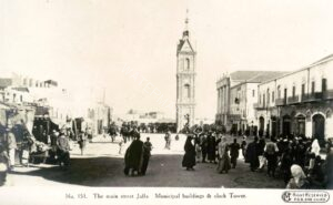 כשני עשורים לאחר שצולמה התמונה הקודמת, ב 16 בנובמבר 1917, כבש הצבא הבריטי את יפו. בגלויה רשמית זו שהופקה (כחלק מסדרה) ע"י הצבא לציון ארועים הקשורים לכיבוש הארץ במלחמת העולם הראשונה (כיבוש צפון הארץ הושלם רק ב 27 בספטמבר 1918), נראה בניין "הסאריה החדשה" (בקצה הימני המרוחק של התמונה), לשעבר מרכז השלטון העות'מני ועתה מרכז השלטון הבריטי. ב 4 בינואר 1948 פוצץ הבנין ע"י אנשי מחתרת לח"י ורק ארבעת עמודי הענק העגולים בחזיתו נותרו במקומם (שחזור חלקי של החזית בוצע שנים מאוחר יותר). המבנה הדו-קומתי המאורך עם כניסותיו המקושתות, בקדמת התמונה בצד ימין, הוא חזיתו של השוק היווני / סוק א-דיר, הוא שוק המנזר (שבבעלות המנזר היווני אורתודוקסי) המודרני והתוסס. האזור כולו המשיך למלא את הפונקציות המרכזיות בחיי העיר גם לאחר תום מלחמת העולם והרחבה הגדולה, ועתה גם מגדל השעון במרכזה, היו ועדיין הינם מסמליה המובהקים.