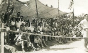 יוםצ ספורט במחנה, קיץ 1946, הדגל הבריטי (ה"יוניון ג'ק") מונף על ראש התורן המאולתר. דב מתחת לסככה, נשען על העמוד התומך מימין (לבוש גופיה). בני משפחות והרבה ילדים צוהלים בין המוזמנים.