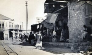 המשכו המדויק של מסלול הרכבת הזעירה (ה"טרזינה") דרך סמטת שער העיר (רח' רוסלאן כיום) ונקודת המפגש שלו עם רחבת "כיכר השעון", מבט ממערב למזרח, צלם לא ידוע. פסי מסילת הרכבת הזעירה שהניחו הבריטים כאמור לאחר כיבוש העיר, נראים כאן בברור. כפי שניתן להתרשם, המסילה (דו-מסלולית !) מתעקלת שמאלה (צפונה) בנקודה זו, ובהמשך, בקצה הצפון-מזרחי של הרחבה, היא תיפנה ימינה (מזרחה) לרחוב בוסטרוס (רזיאל כיום), אל עבר שדרות קינג ג'ורג' (שד' ירושלים כיום), ודרך שכונת מנשיה תעשה דרכה אל תחנת הרכבת. 
החנויות של בעלי-העסקים סיקסיק ומויאל בימין התמונה, קיר האבנים והקשת במרכזו קיים עד היום (רח' רוסלאן מס' 6) וחזית "השוק היווני" הדו-קומתי בקדמת התמונה. עמודי החשמל, בתמונה זו כבקודמתה, מהווים רמז נוסף לזמן בו צולמו השתיים, שכן, בשנים הללו בדיוק (אמצע שנות ה 20 של המאה העשרים), יפו (כמו גם תל אביב) חוברה לרשת החשמל. באותן שנים גם תוציא הרכבת הזעירה את ימיה ופסיה יפורקו בהדרגה.